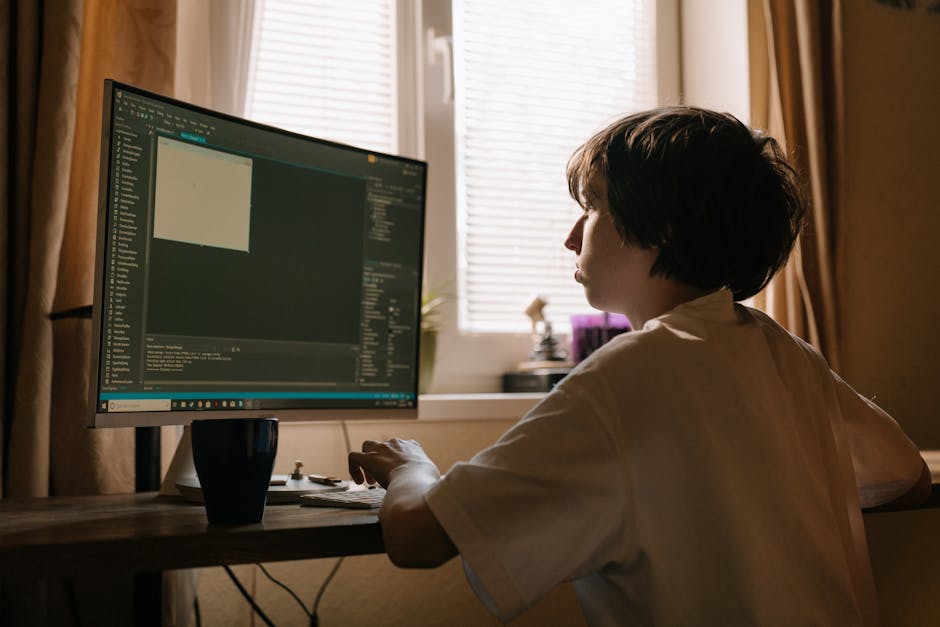 A person with short dark hair and wearing a white shirt is seated at a wooden desk in a room with natural light coming through partially open blinds. They are working on a desktop computer with a larg