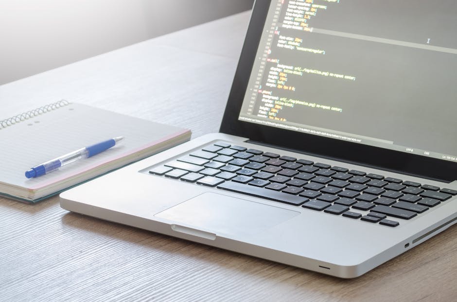 A close-up view of a silver laptop displaying lines of colorful code on a dark background, placed on a light wooden desk. Next to the laptop is an open spiral-bound notebook with a white cover, accomp