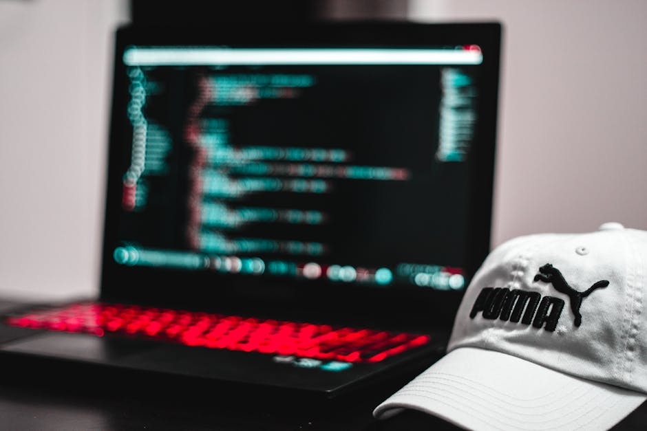 A close-up view of a silver laptop displaying lines of colorful code on a dark background, placed on a light wooden desk. Next to the laptop is an open spiral-bound notebook with a white cover, accomp